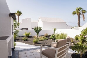 Een terras met een witte stoel, diverse planten en uitzicht op witte gebouwen en palmbomen.