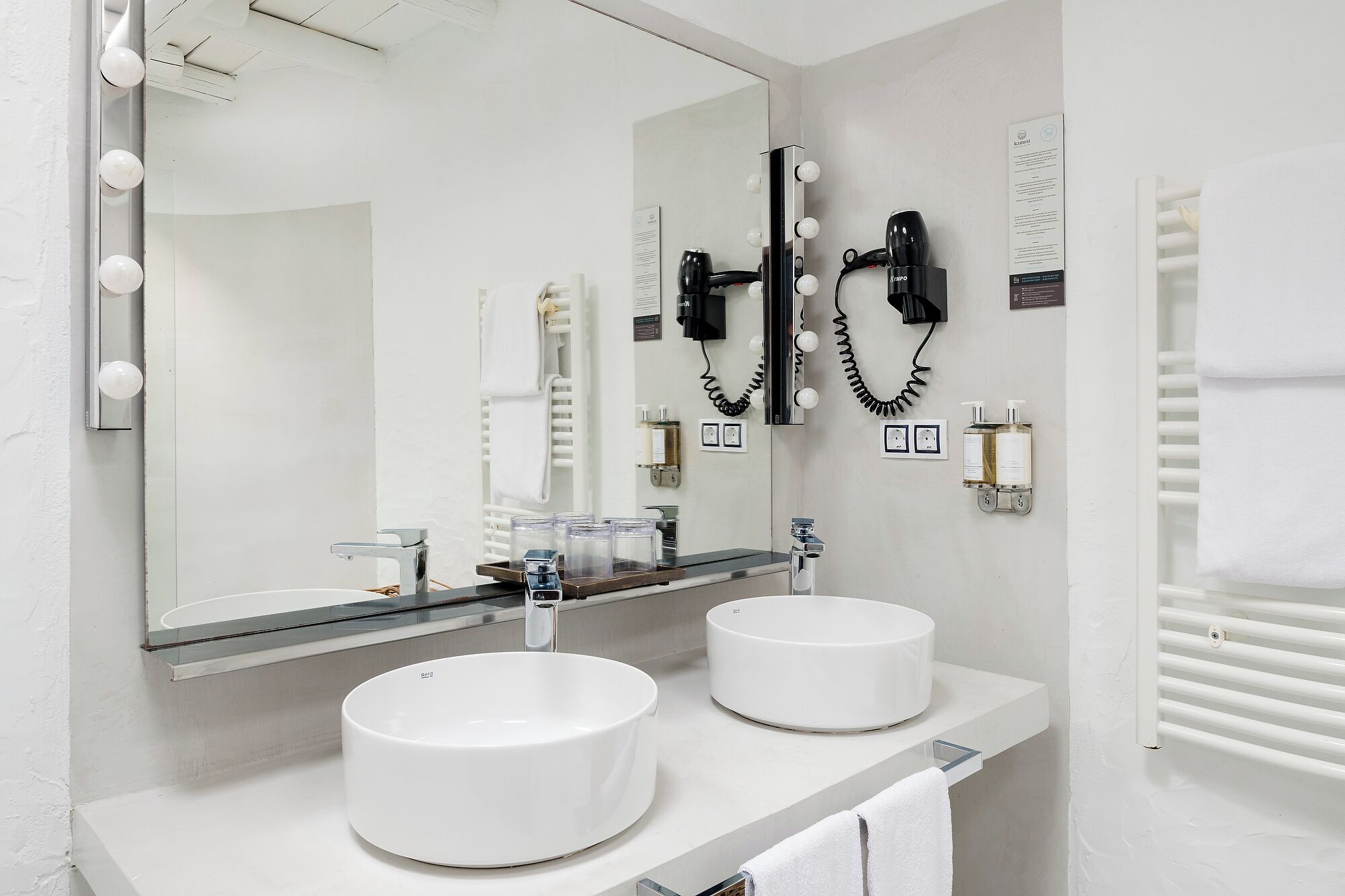 A modern bathroom with two round sinks, a large mirror with lights, a hairdryer, and a towel rack.