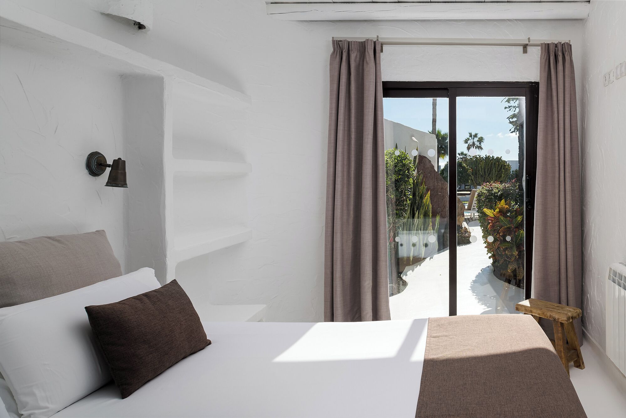 Un dormitorio con una cama, una mesita de noche, una lámpara y vistas a un jardín a través de un ventanal corredero.