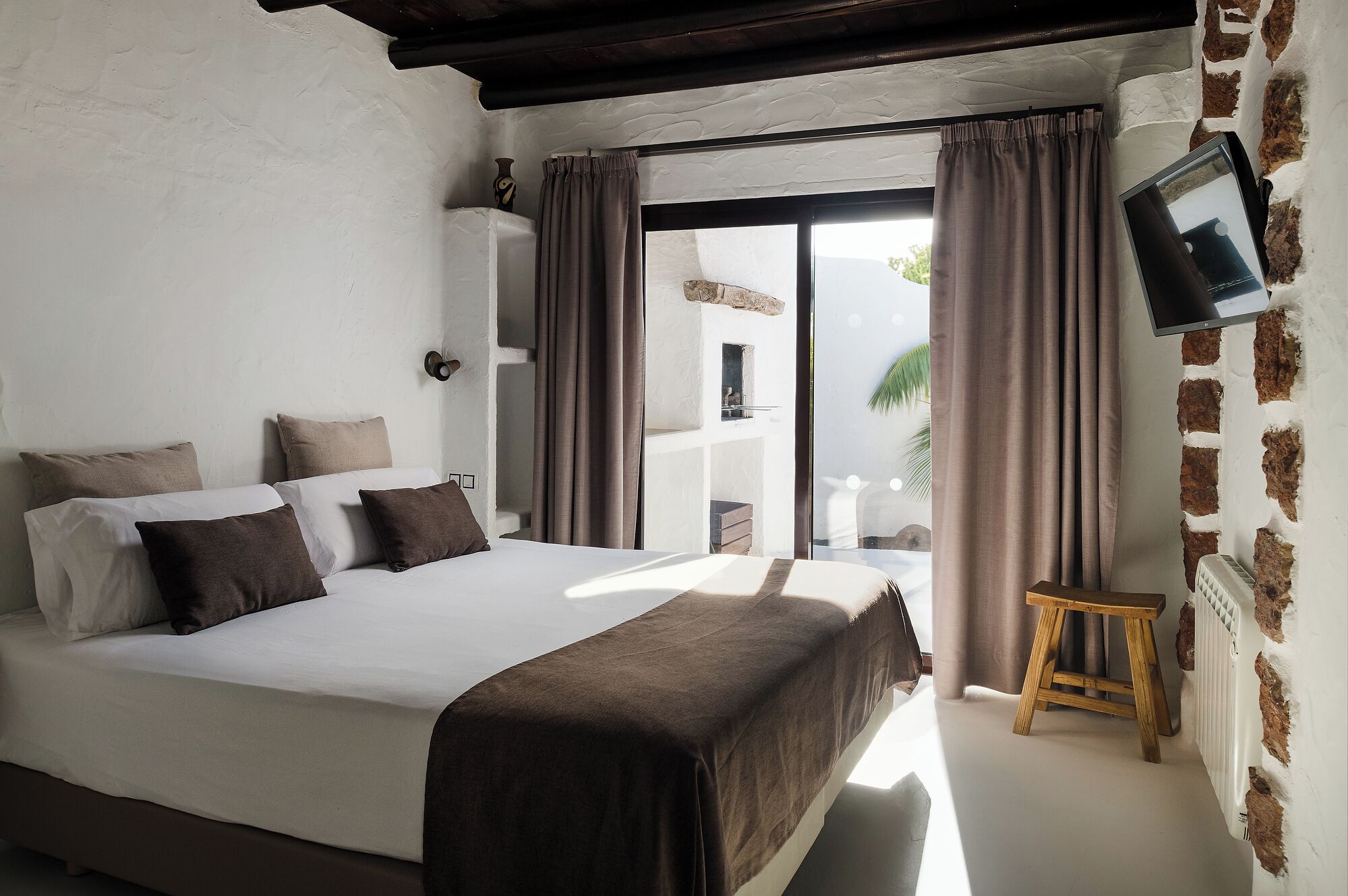 A bedroom with a large bed, a wooden stool, and a TV mounted on the wall.