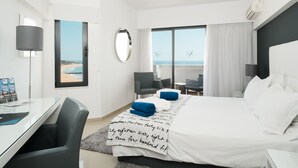 Habitación de hotel con cama, escritorio, silla, ventana grande con vista a la playa y un equipo de aire acondicionado.