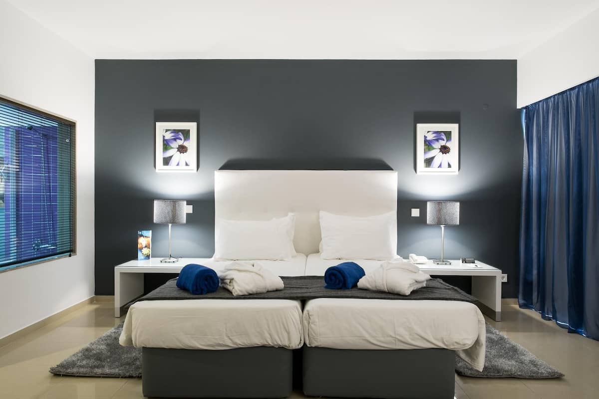 A modern hotel room with a large bed, two bedside tables, a desk, and a window with blinds.