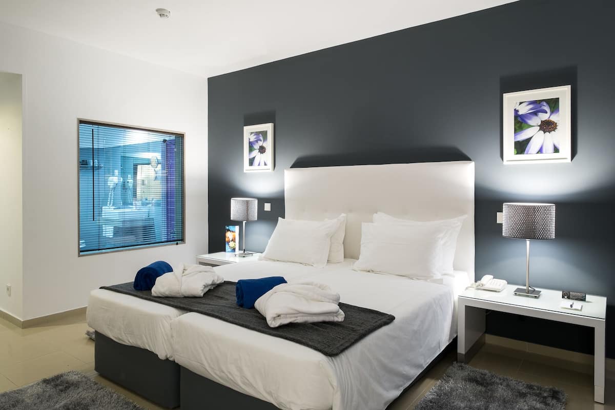 A modern hotel room with a large bed, a bedside table, a nightstand, a window with blinds, and a wall-mounted picture.