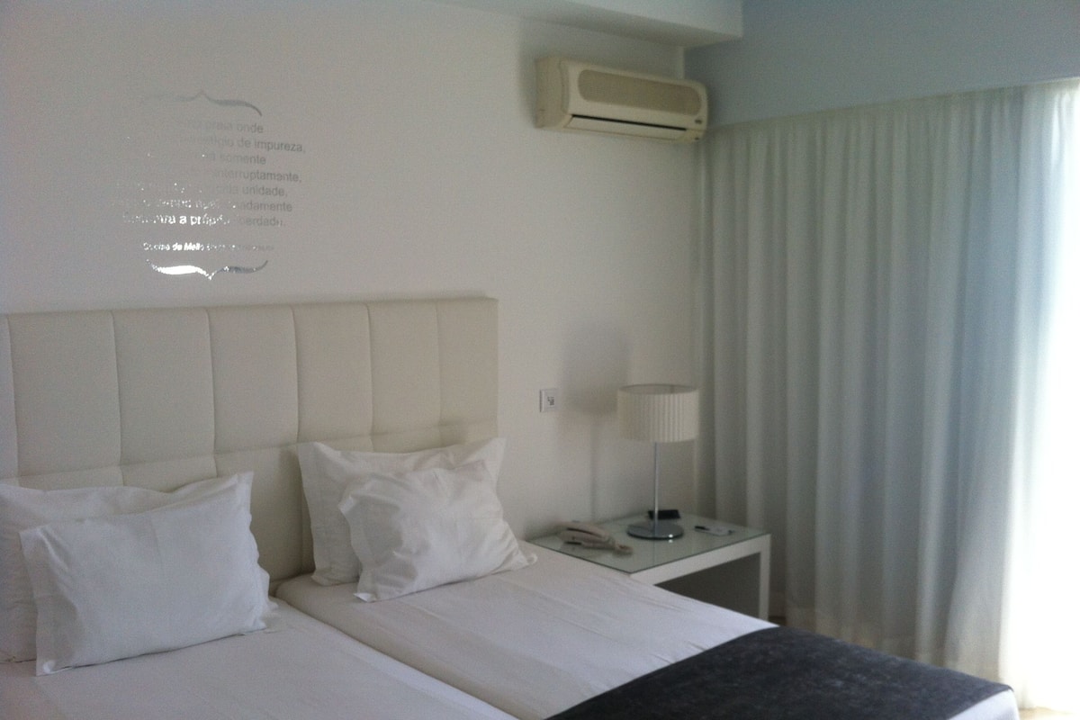 A hotel room with two beds, a bedside table with a lamp, and a window with curtains.