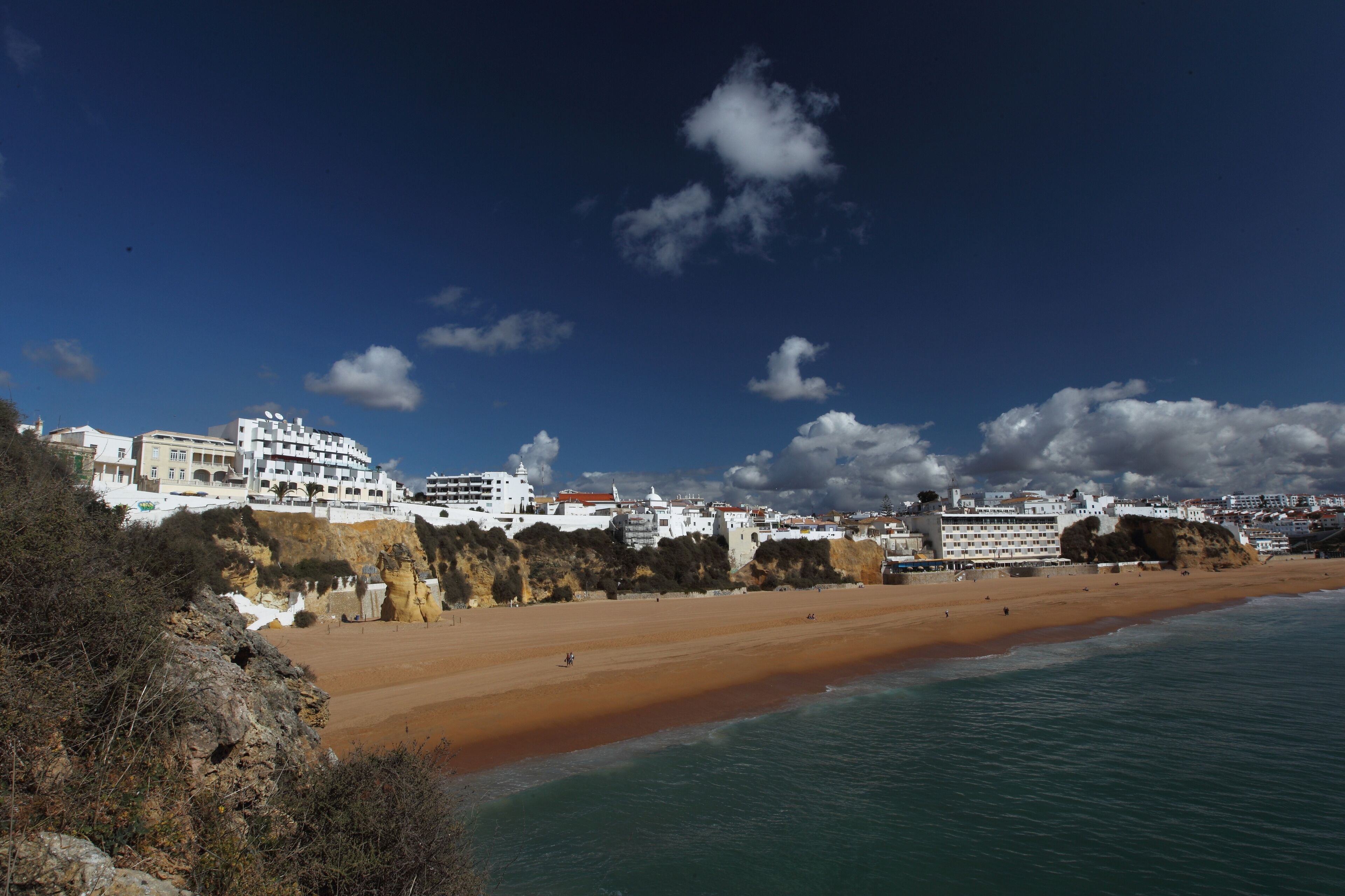 A coastal area with a sandy beach, rocky cliffs, and white buildings with balconies.