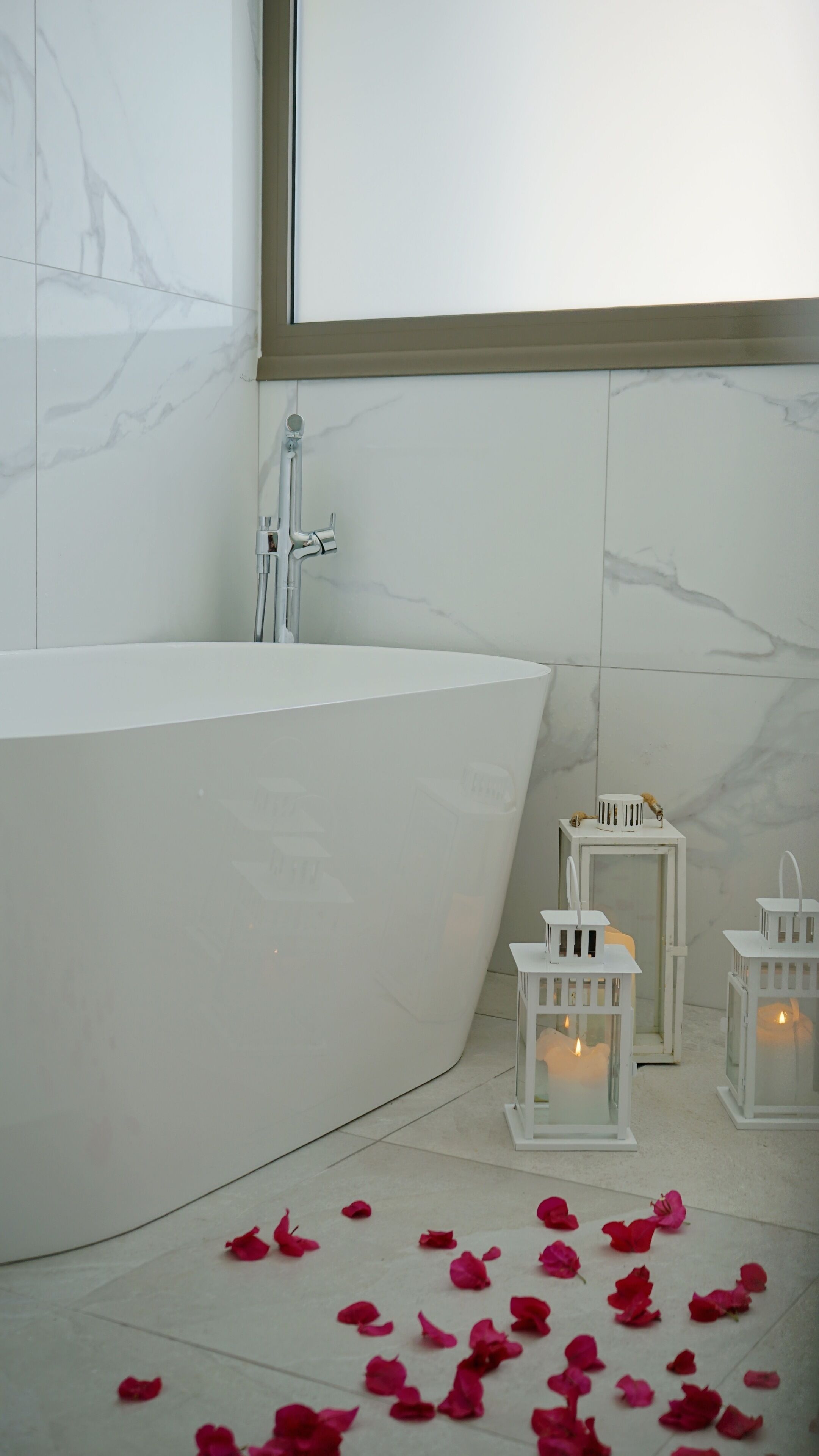 A bathroom with a freestanding bathtub, white marble tiles, and two lit white lanterns on the floor.