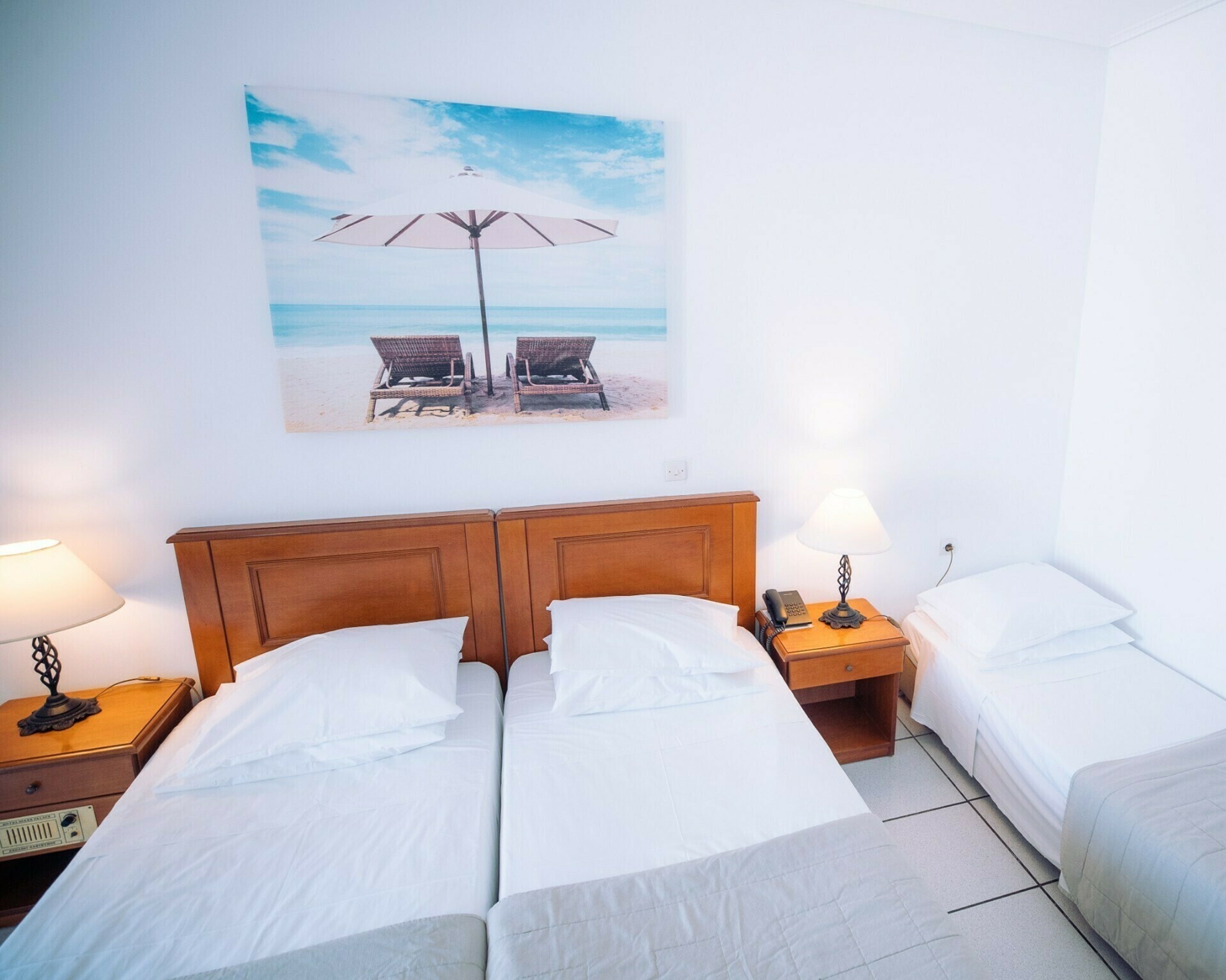 A hotel room with two single beds, a wooden headboard, a nightstand with a lamp, a painting of beach chairs, and a beach scene.