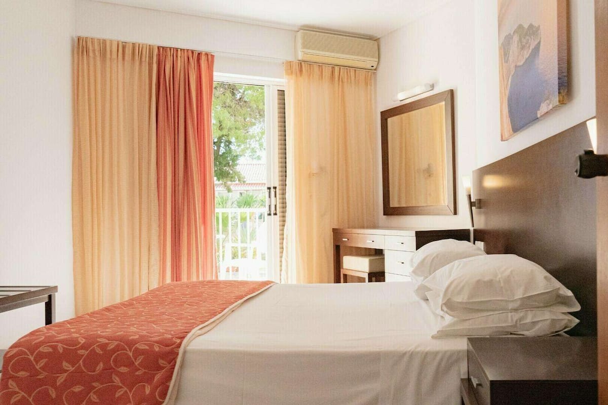 Une chambre d’hôtel comprenant un lit, un bureau, une chaise, une commode, un miroir et une fenêtre avec des rideaux.