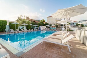Una piscina con lettini e ombrelloni, immersa nel verde e circondata da edifici.