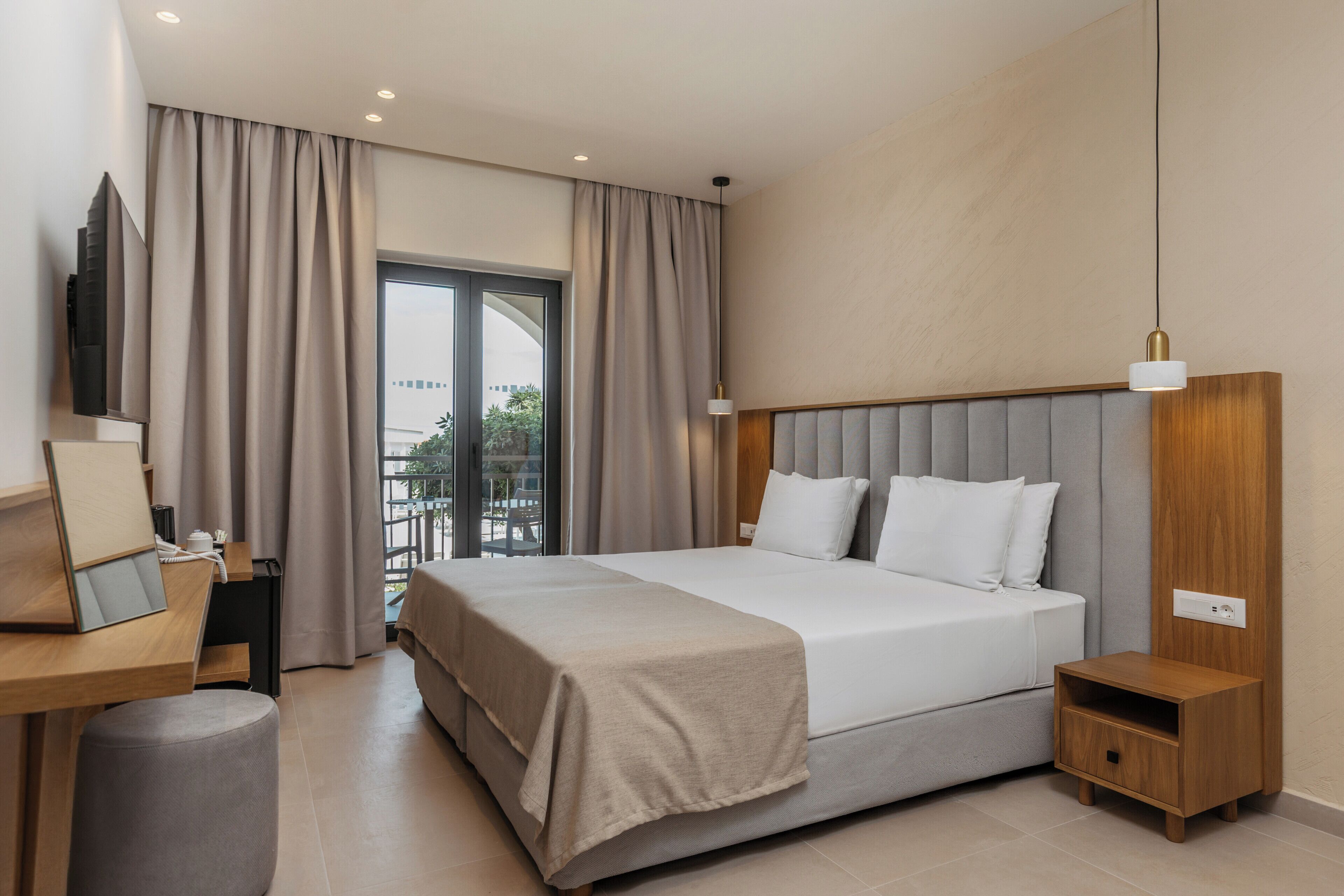 A modern hotel room with a large bed, a desk with a computer, a small bedside table, and a large window with curtains.