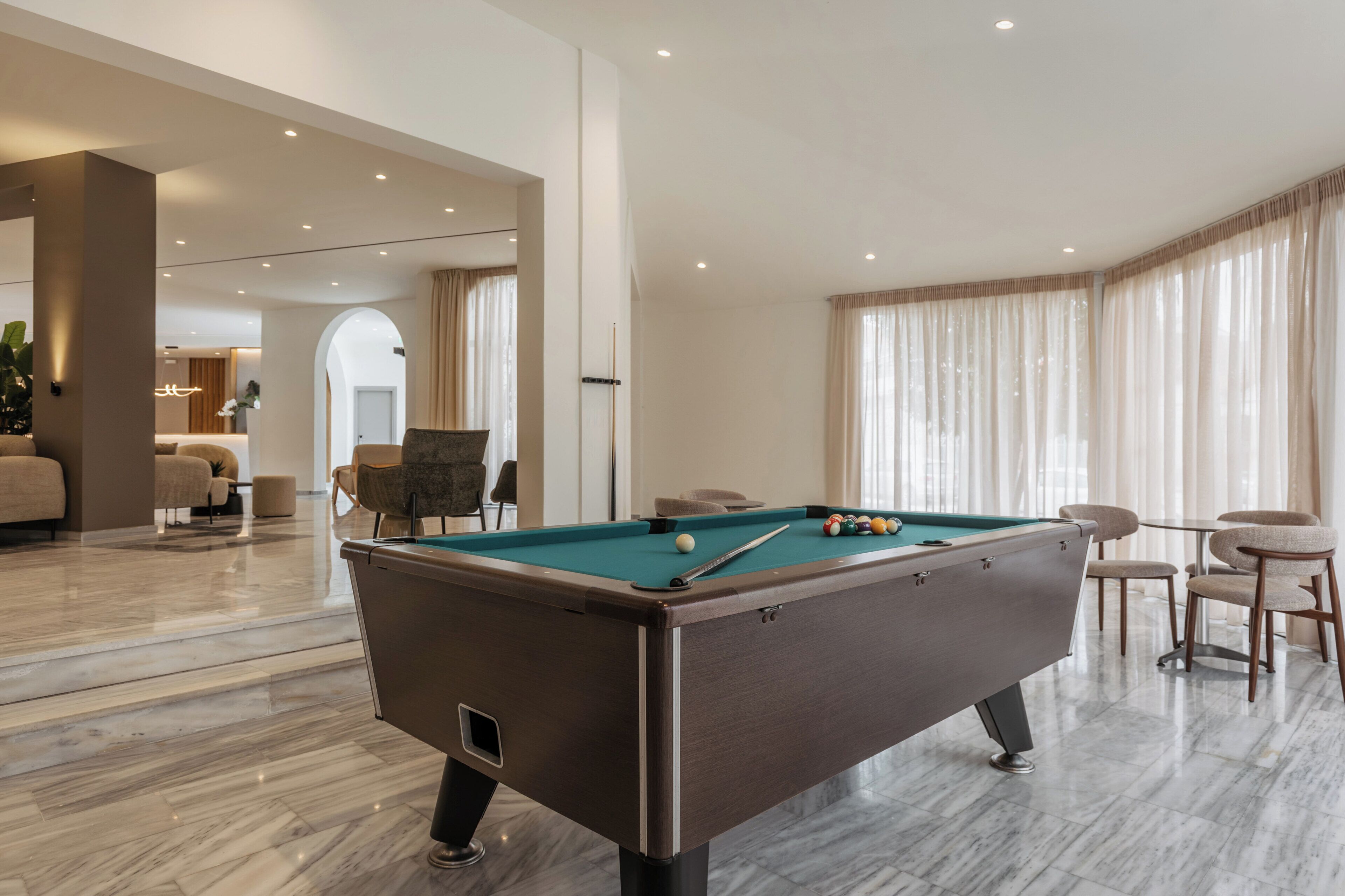 Een biljart tafel in een goed verlichte kamer met een marmeren vloer, een zithoek met stoelen en een groot raam met gordijnen.