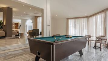 A pool table in a well-lit room with a marble floor, a seating area with chairs, and a large window with curtains.