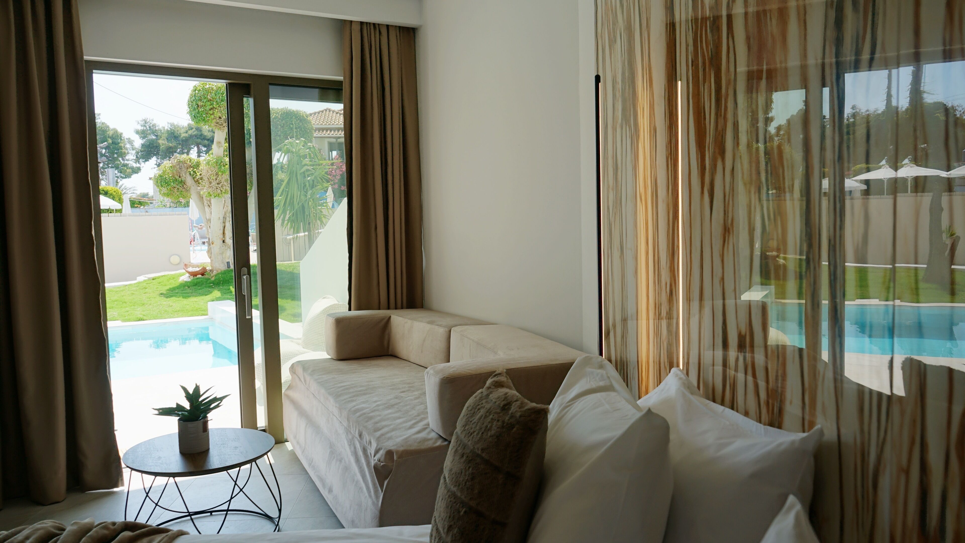 A modern hotel room with a large sofa, a small round table, and a view of a swimming pool and garden.