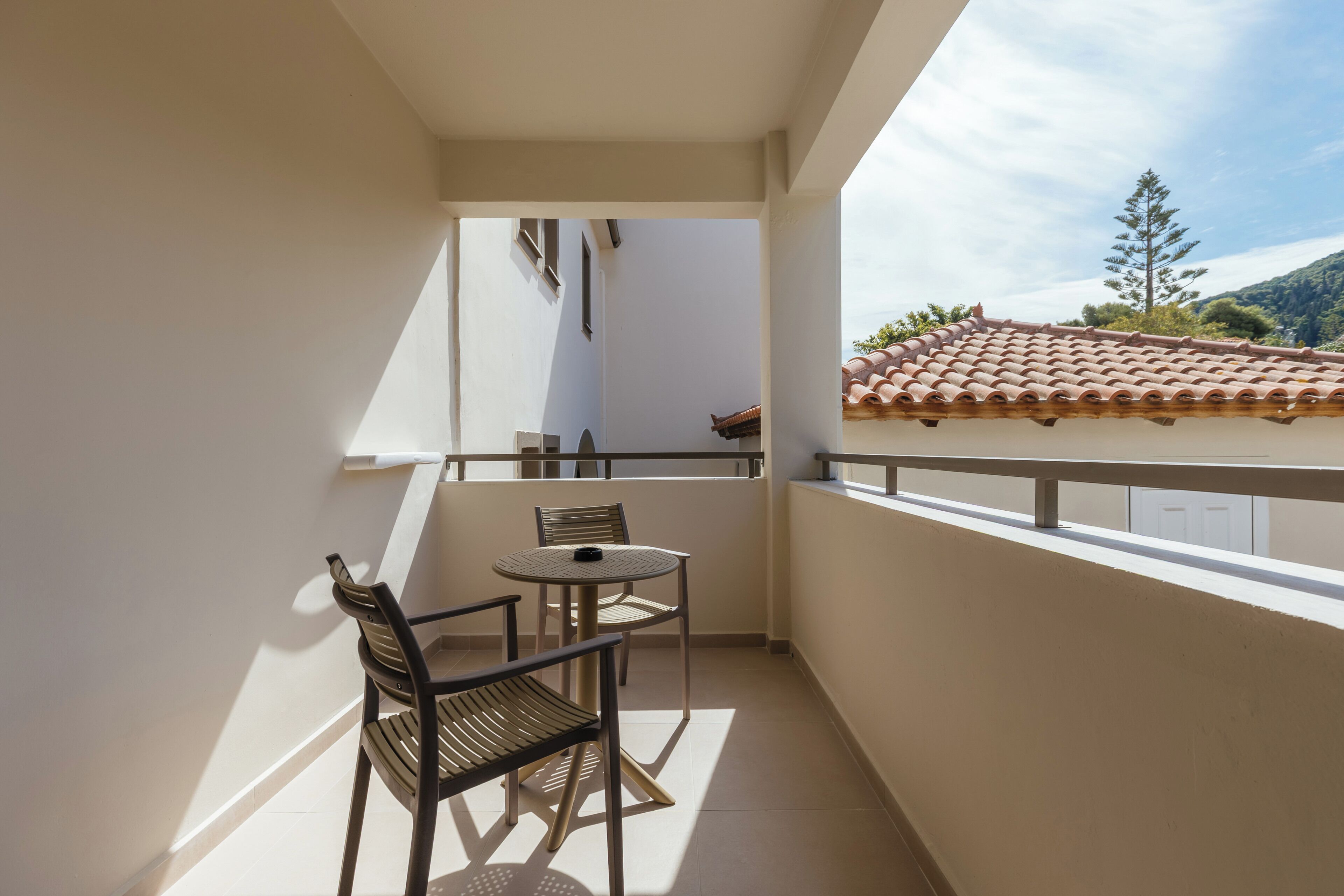 A balcony with a table and two chairs, a white wall, and a view of a building with a tiled roof.