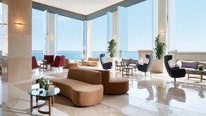 A spacious lobby with a modern design, featuring a mix of seating options and large windows offering a view of the sea. - Ikos Oceania - All Inclusive (Nea Propontida)