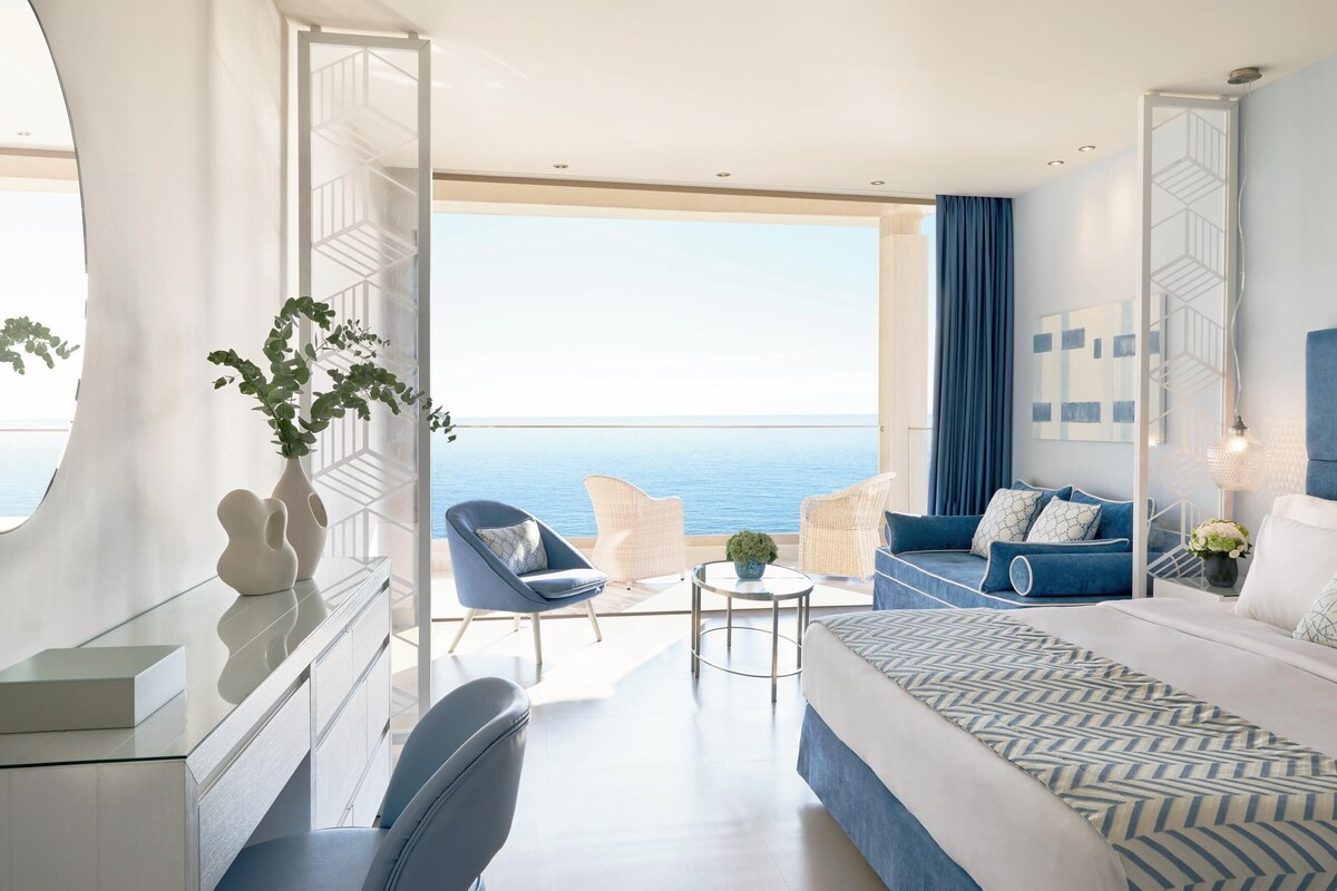 A modern hotel room with a large balcony, a bed, a desk, and seating area.