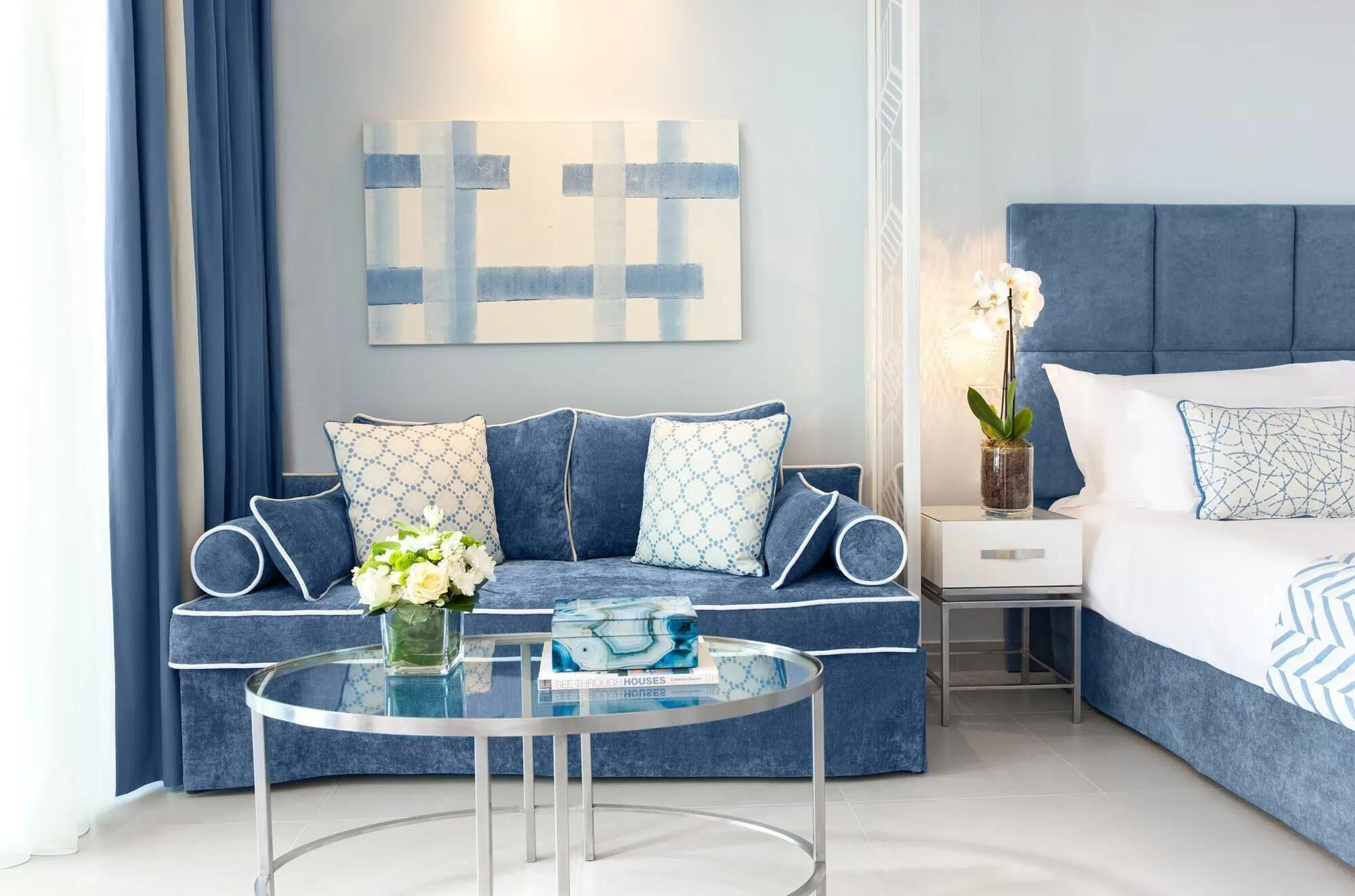 A modern bedroom with a blue sofa, a glass coffee table, a bedside table with a lamp, and a bed with a blue headboard.