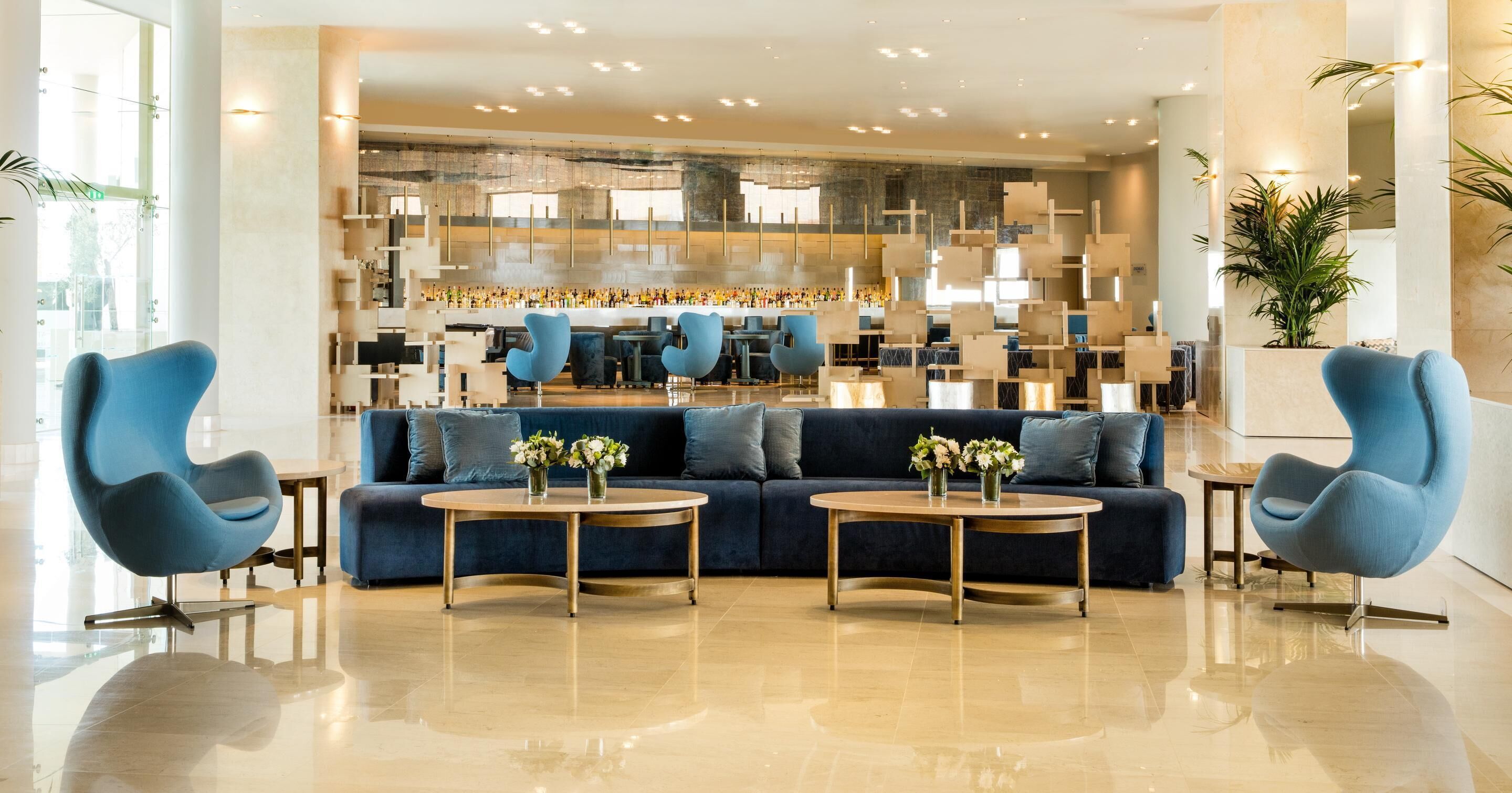 A modern lobby with a blue sofa, two round tables with vases, and a bar area in the background.