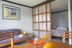 A room with a sofa, a table with a fruit juicer and glasses, a bed with a colorful blanket, and a sliding door. - Sowell LES Cavales (Carcans)