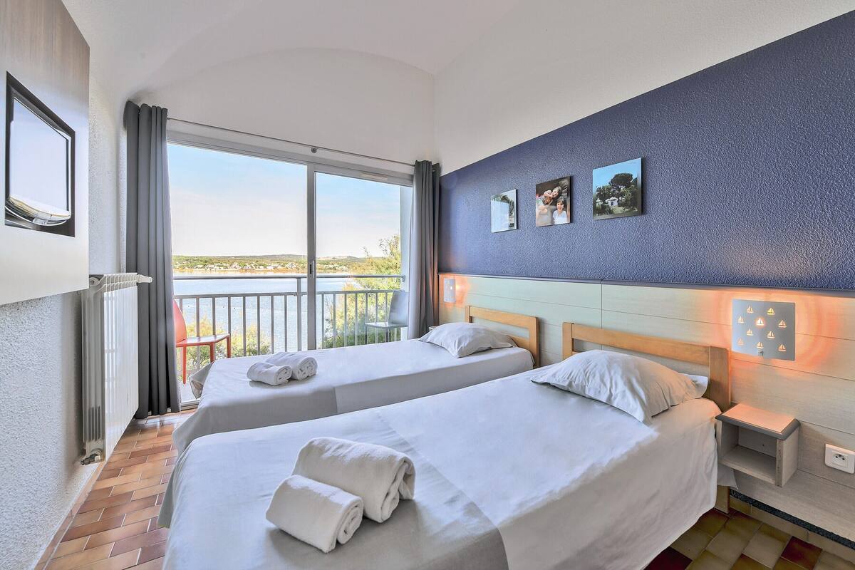 A hotel room with two beds, a view of the sea, a balcony, and a blue textured wall with pictures.