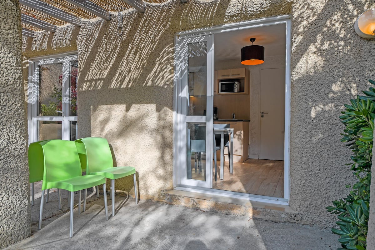A cozy outdoor seating area with green chairs and a table, adjacent to a modern kitchen with a microwave and a hanging light fixture.