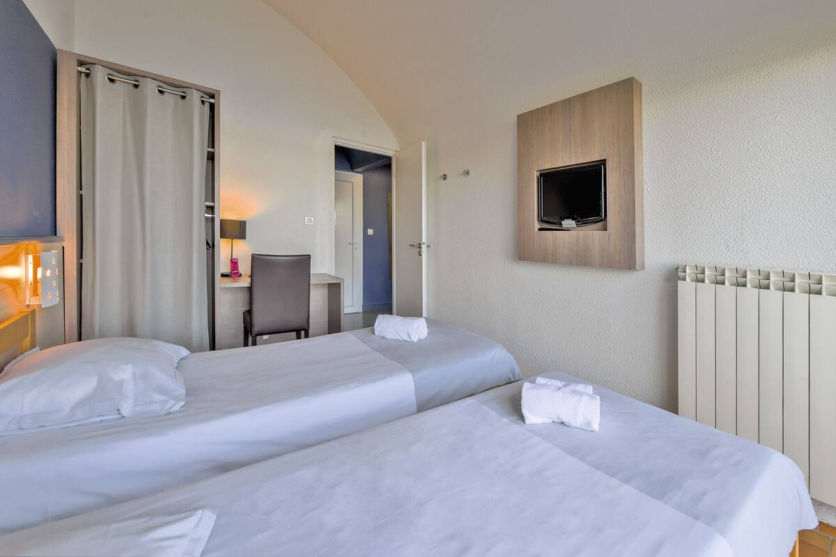 A hotel room with two beds, a desk, a chair, a TV mounted on the wall, and a radiator.