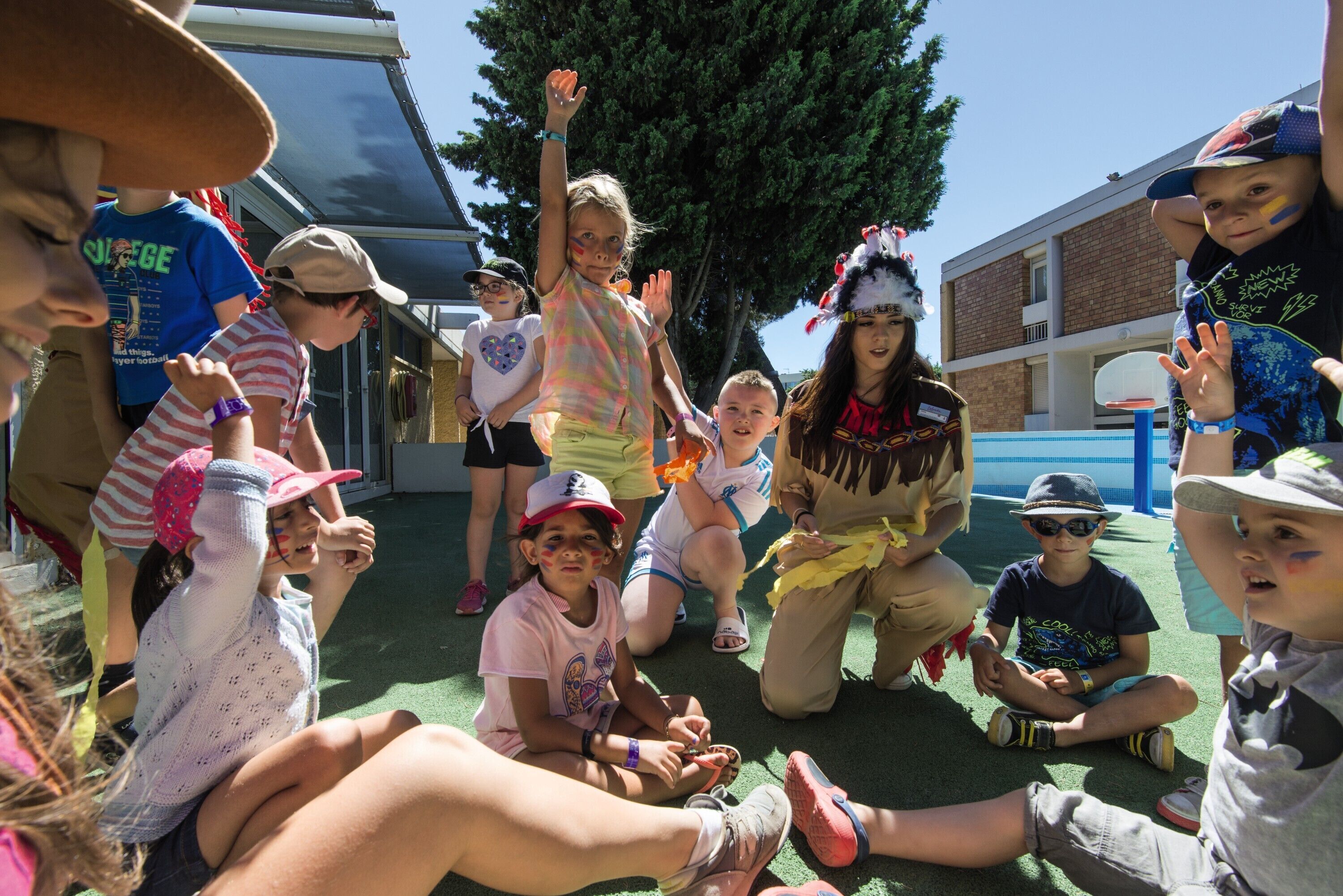 Un groupe d’enfants assis par terre, à l’extérieur, dont certains portent des chapeaux et des vêtements décontractés.