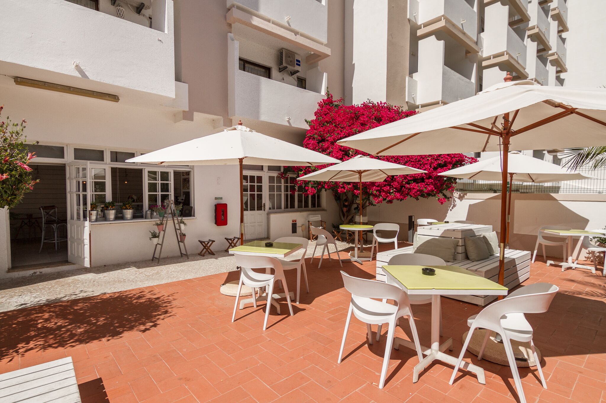 An outdoor patio with white tables and chairs, surrounded by white buildings and red flowers.