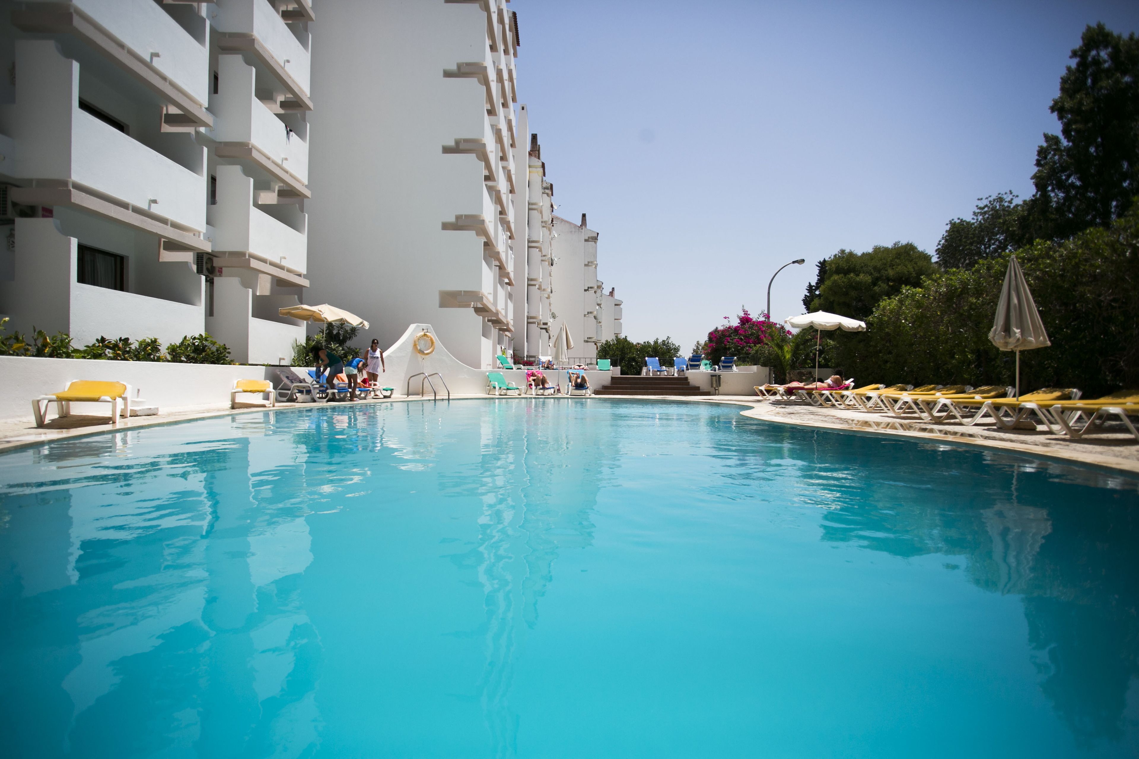 Una piscina con tumbonas y sombrillas, rodeada por un edificio blanco con balcones.