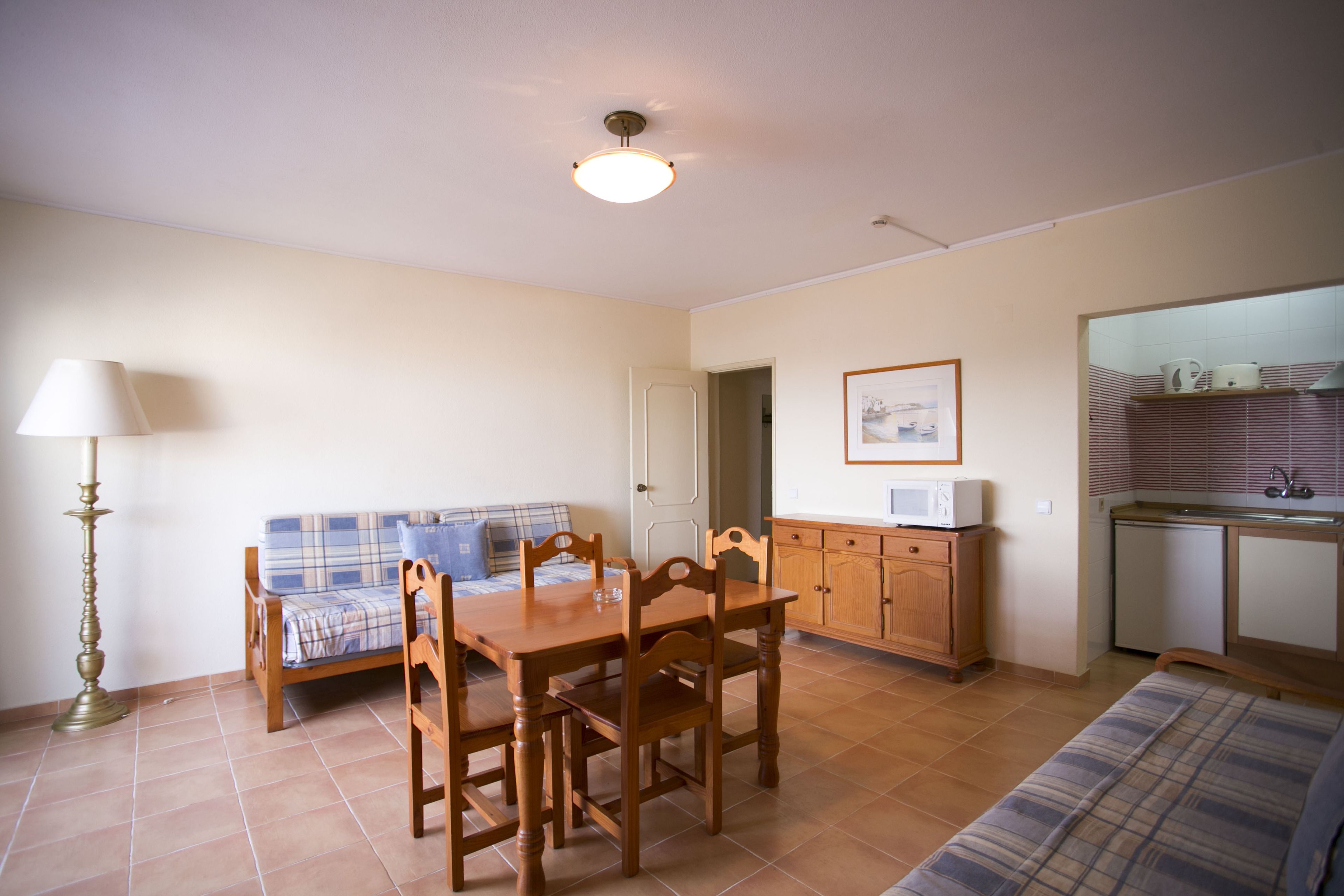 A compact living space with a dining table, wooden chairs, a sofa, a lamp, a microwave, and a small kitchenette.