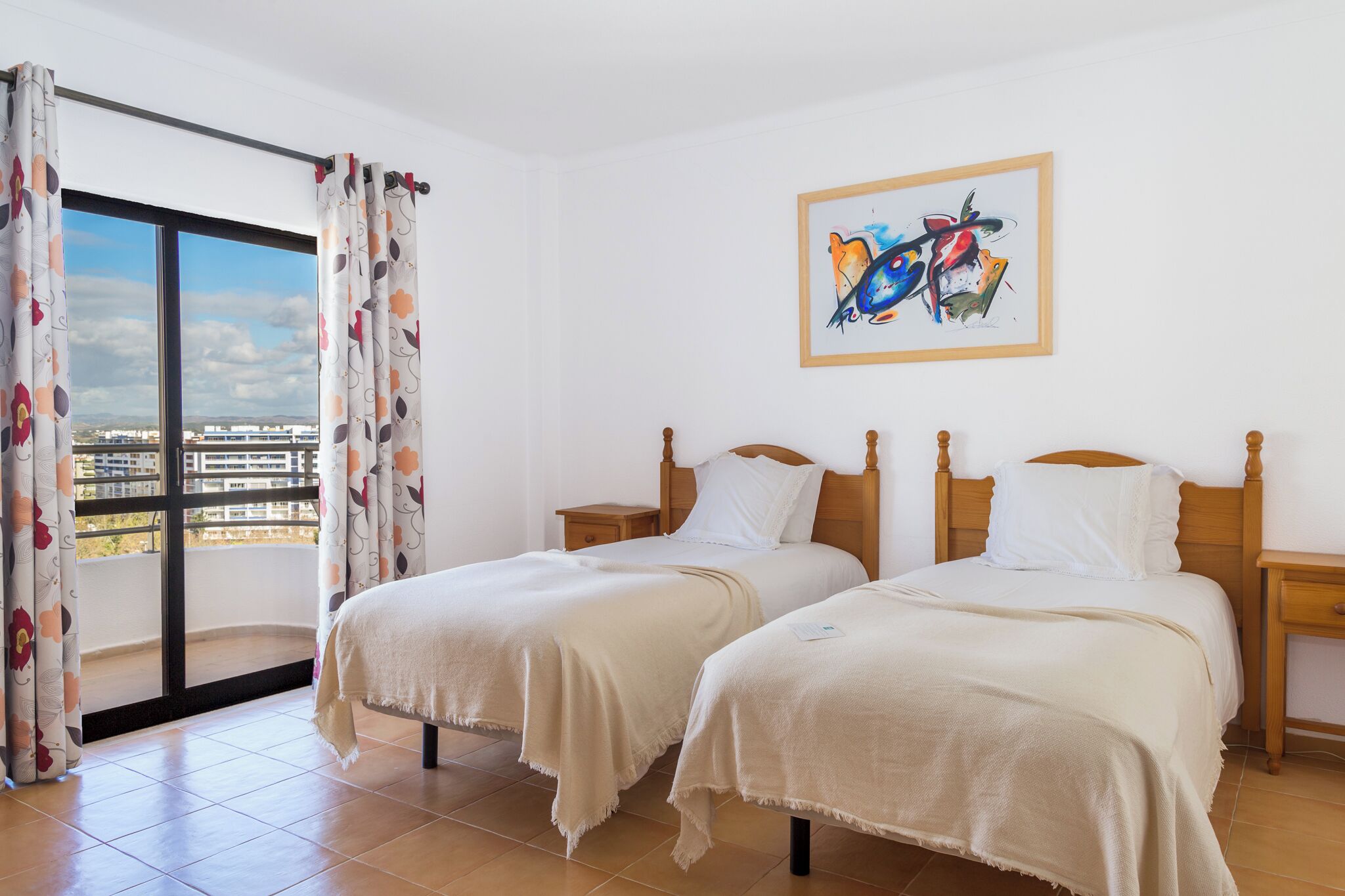 Een hotelkamer met twee bedden, een balkon met gordijnen, een schilderij aan de muur en uitzicht op de stad.