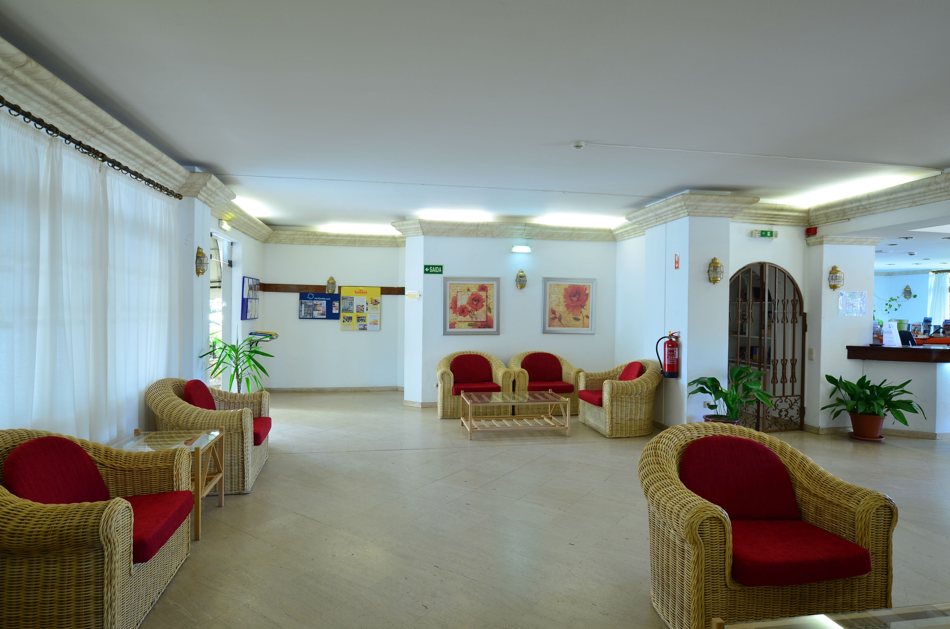 A lobby with wicker furniture, red cushions, and potted plants.