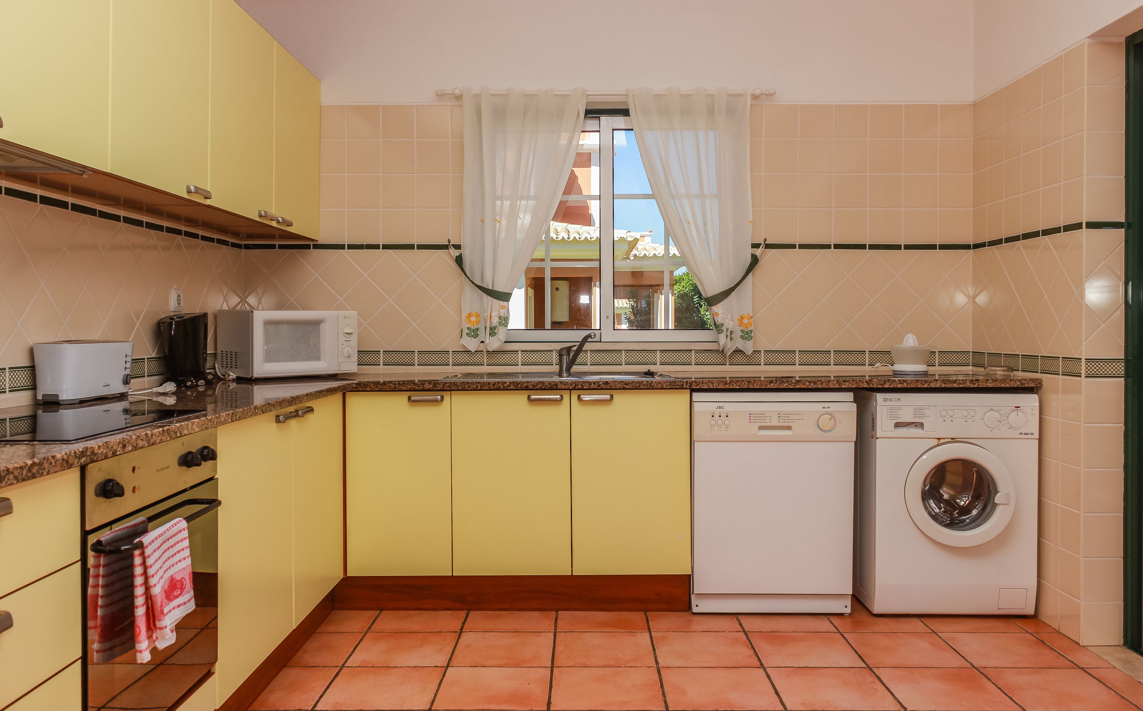Una cocina con armarios amarillos, microondas, cafetera, lavadora y ventana con cortinas.