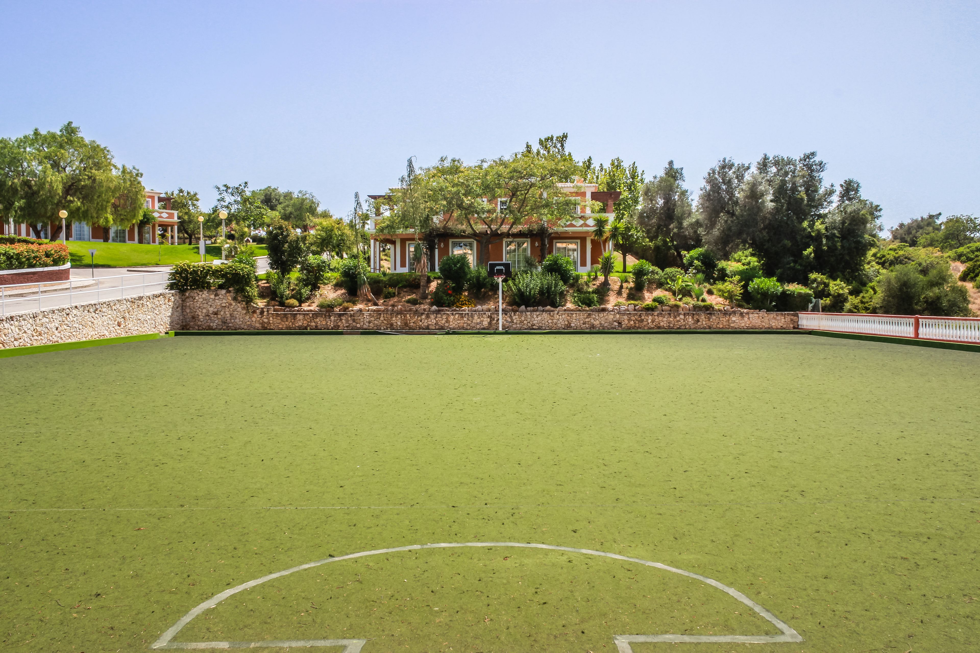 Un terrain de sport vert, doté d’un panier de basketball, entouré d’un mur de pierre et d’arbres.