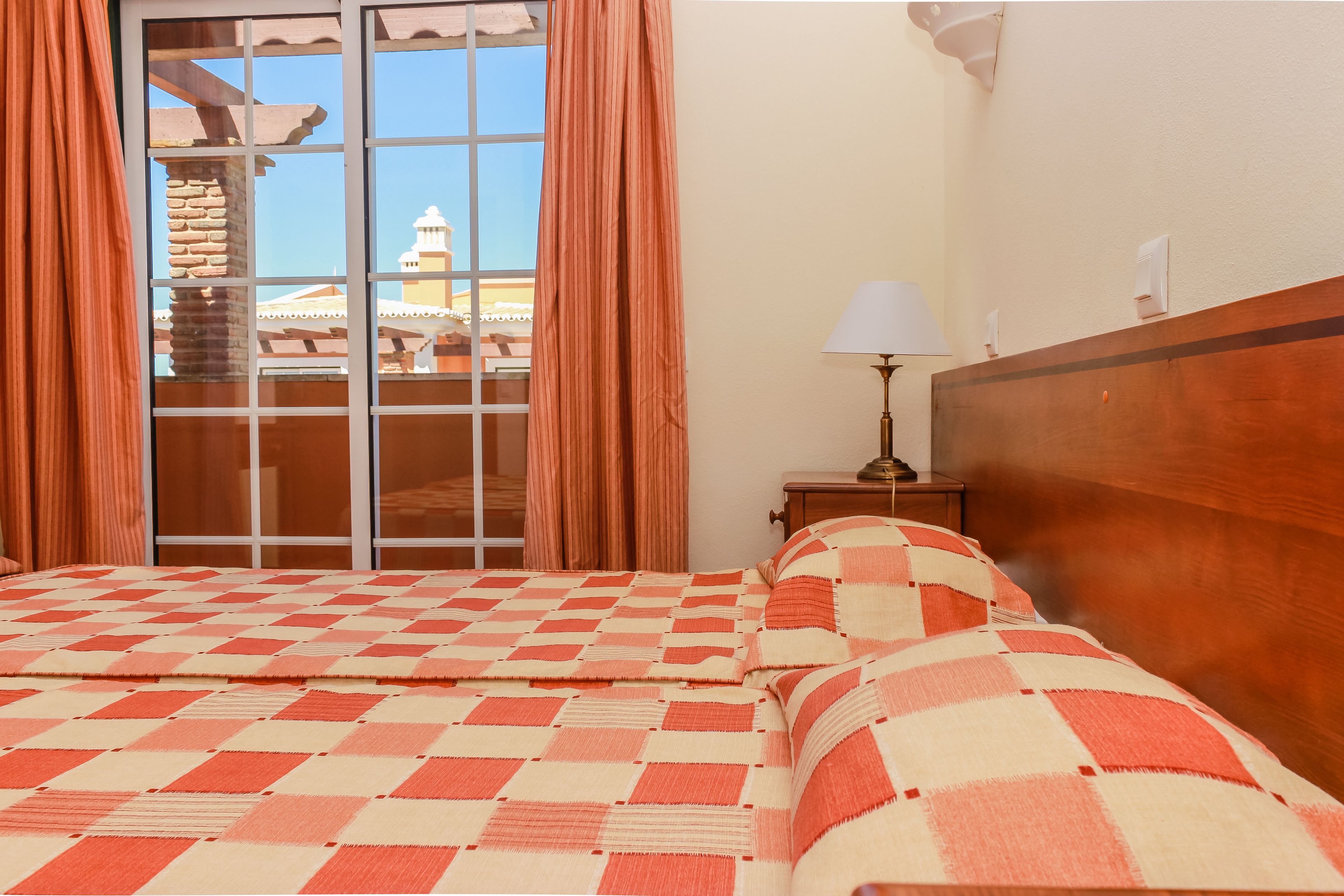 A hotel room with a checkered bedspread, a wooden headboard, a nightstand with a lamp, and a view of a building through a window with curtains.