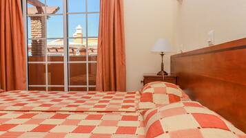 A hotel room with a checkered bedspread, a wooden headboard, a nightstand with a lamp, and a view of a building through a window with curtains.