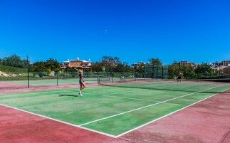 Una cancha de tenis con una red, rodeada por una cerca, y edificios al fondo.