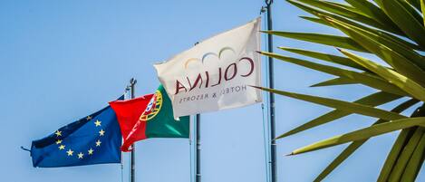 Two flags, one of the European Union and one of Portugal, flying on flagpoles against a clear blue sky.