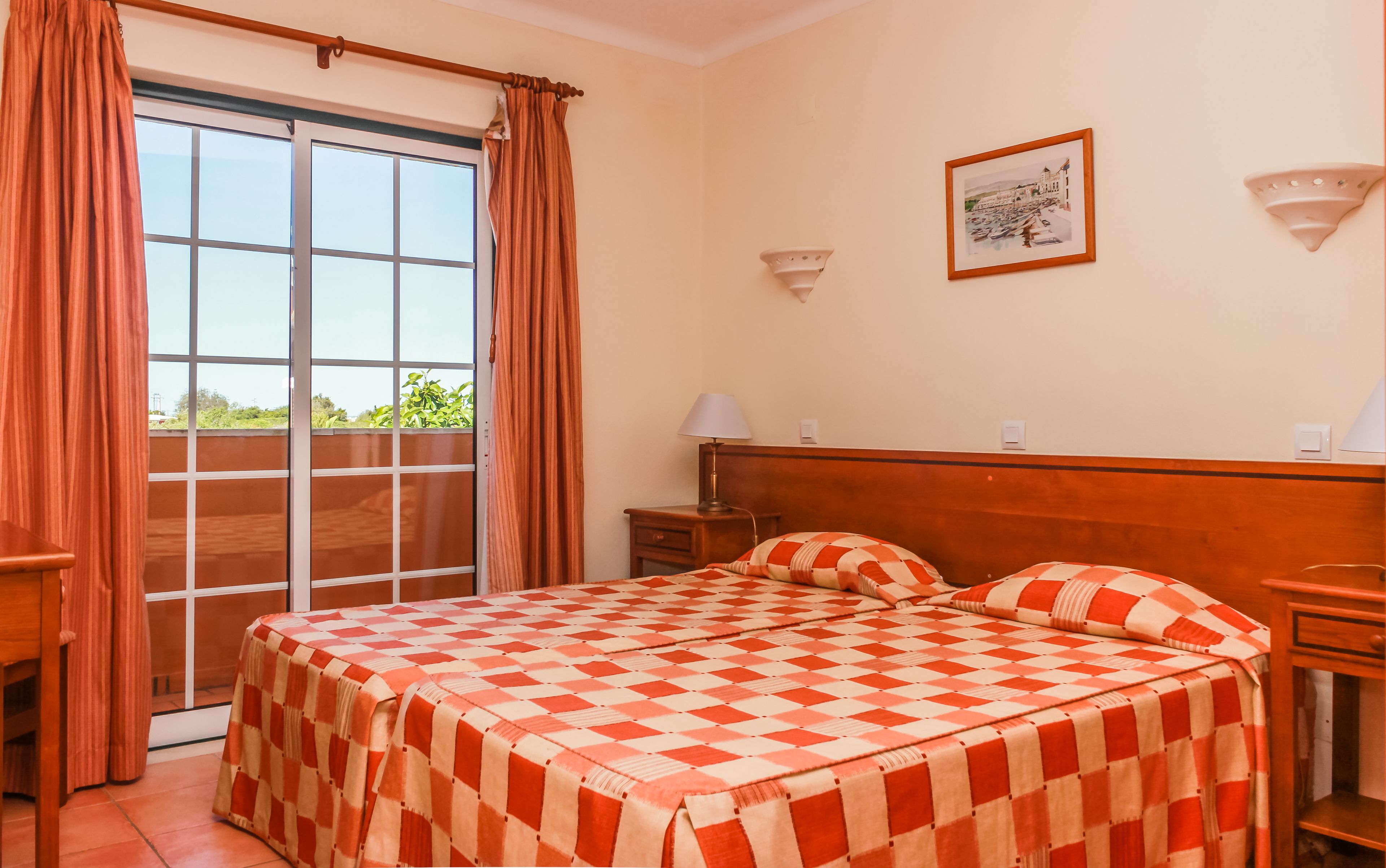 A hotel room with a checkered bedspread, a wooden headboard, a nightstand with a lamp, and a window with curtains.