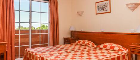 A hotel room with a checkered bedspread, a wooden headboard, a nightstand with a lamp, and a window with curtains.