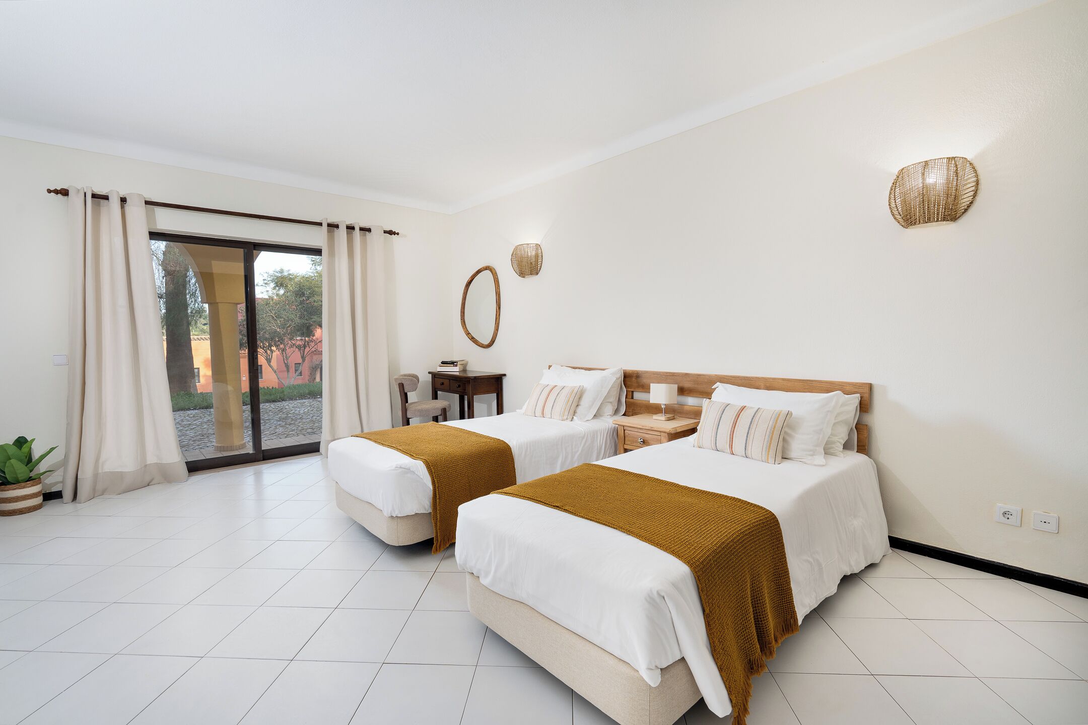 Habitación de hotel con dos camas, una puerta corrediza de vidrio, una mesita y una planta en maceta.