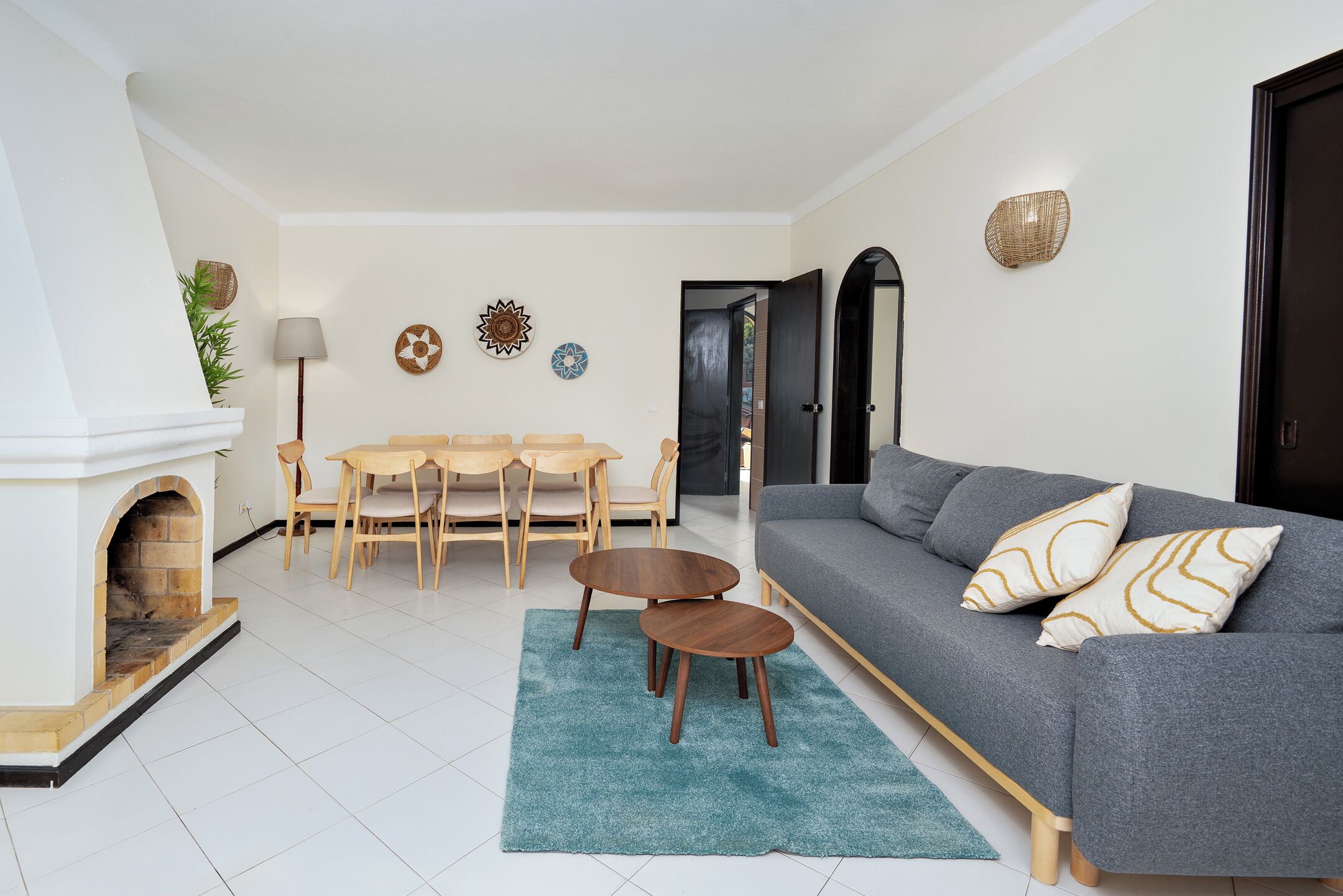 Een woonkamer met een grijze bank, een open haard, een eettafel met stoelen en een blauw vloerkleed.