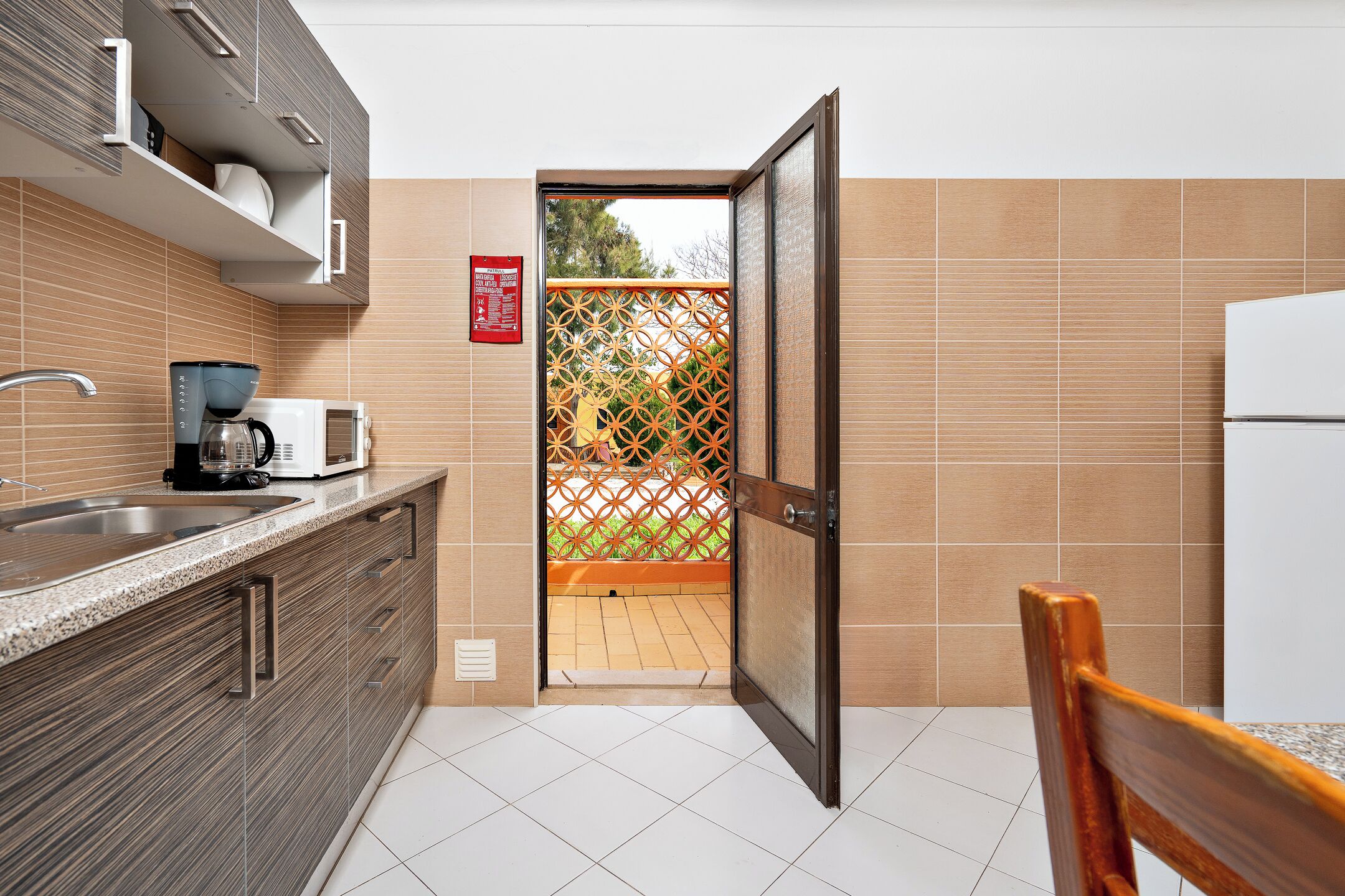A kitchen with a built-in microwave, coffee maker, and a sink. There's a wooden chair and a door leading outside.
