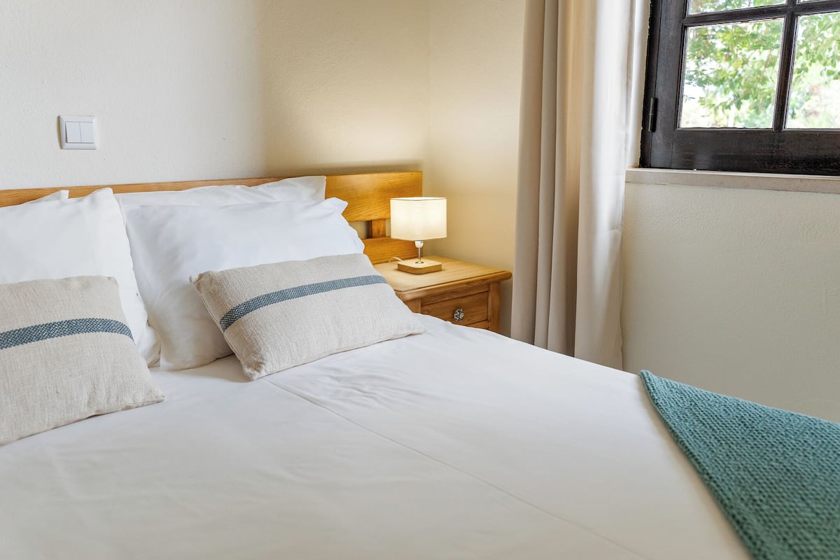 Un lit bien fait, avec du linge de lit blanc et deux oreillers, une table de chevet avec une lampe et une fenêtre avec des rideaux.