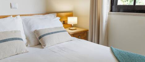 Una cama bien tendida con sábanas blancas y dos almohadas, una mesita de noche con una lámpara, y una ventana con cortinas.