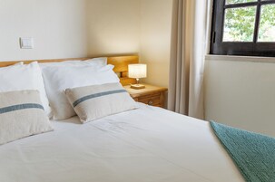 Un lit bien fait, avec du linge de lit blanc et deux oreillers, une table de chevet avec une lampe et une fenêtre avec des rideaux.