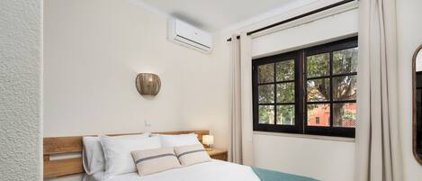 Una cama bien tendida con sábanas blancas y una manta turquesa, una lámpara de noche y una ventana con cortinas.