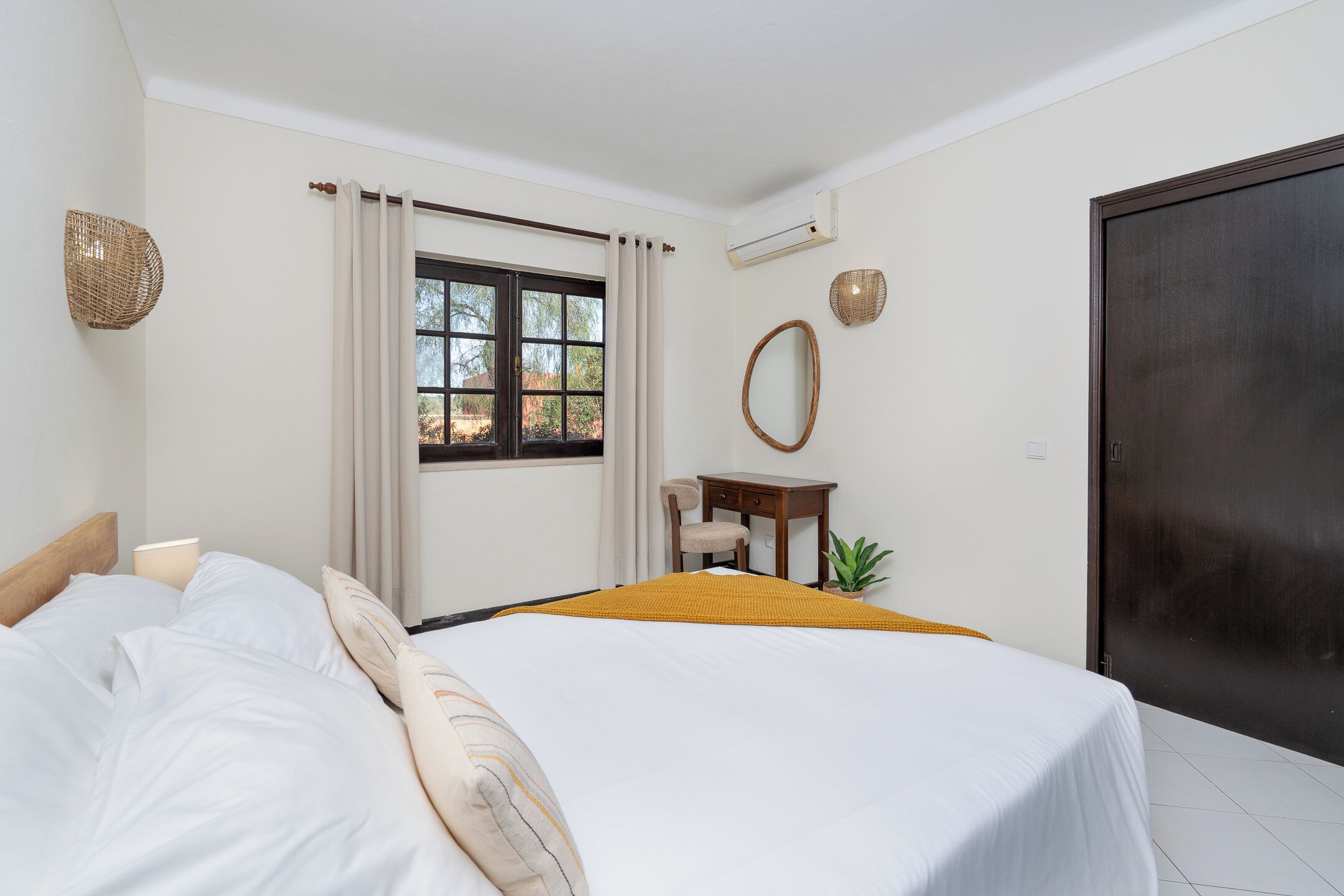 A bedroom with a bed, a desk, a chair, a window with curtains, a wall-mounted mirror, and a door.