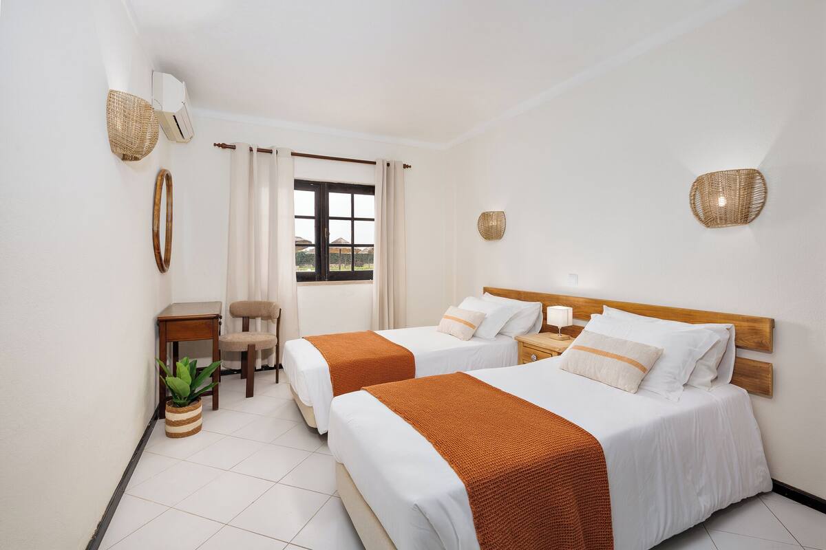 Une chambre d’hôtel avec deux lits, une petite table, une chaise, une plante en pot et une fenêtre avec des rideaux.