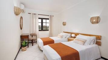 A hotel room with two beds, a small table, a chair, a potted plant, and a window with curtains.
